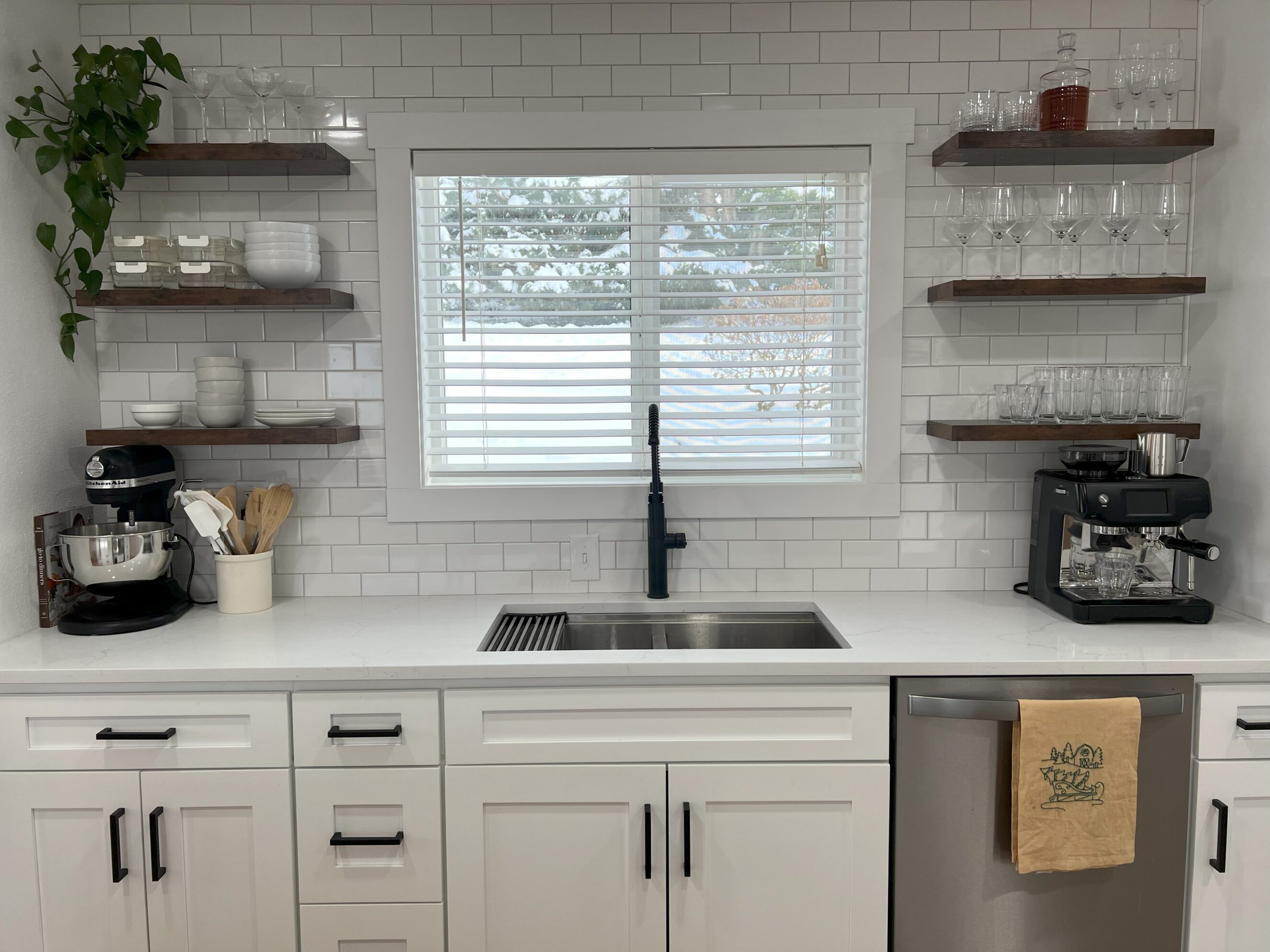 Bright modern kitchen showroom scene featuring white subway tile backsplash, white quartz countertop, undermount stainless sink with matte black faucet beneath a window with blinds, white shaker cabinets with black hardware, floating dark wood shelves displaying dishes, glassware and plants, stainless dishwasher with a hanging tea towel, stand mixer and espresso machine on the counter — showcasing tile, countertops, cabinets, and showroom design.