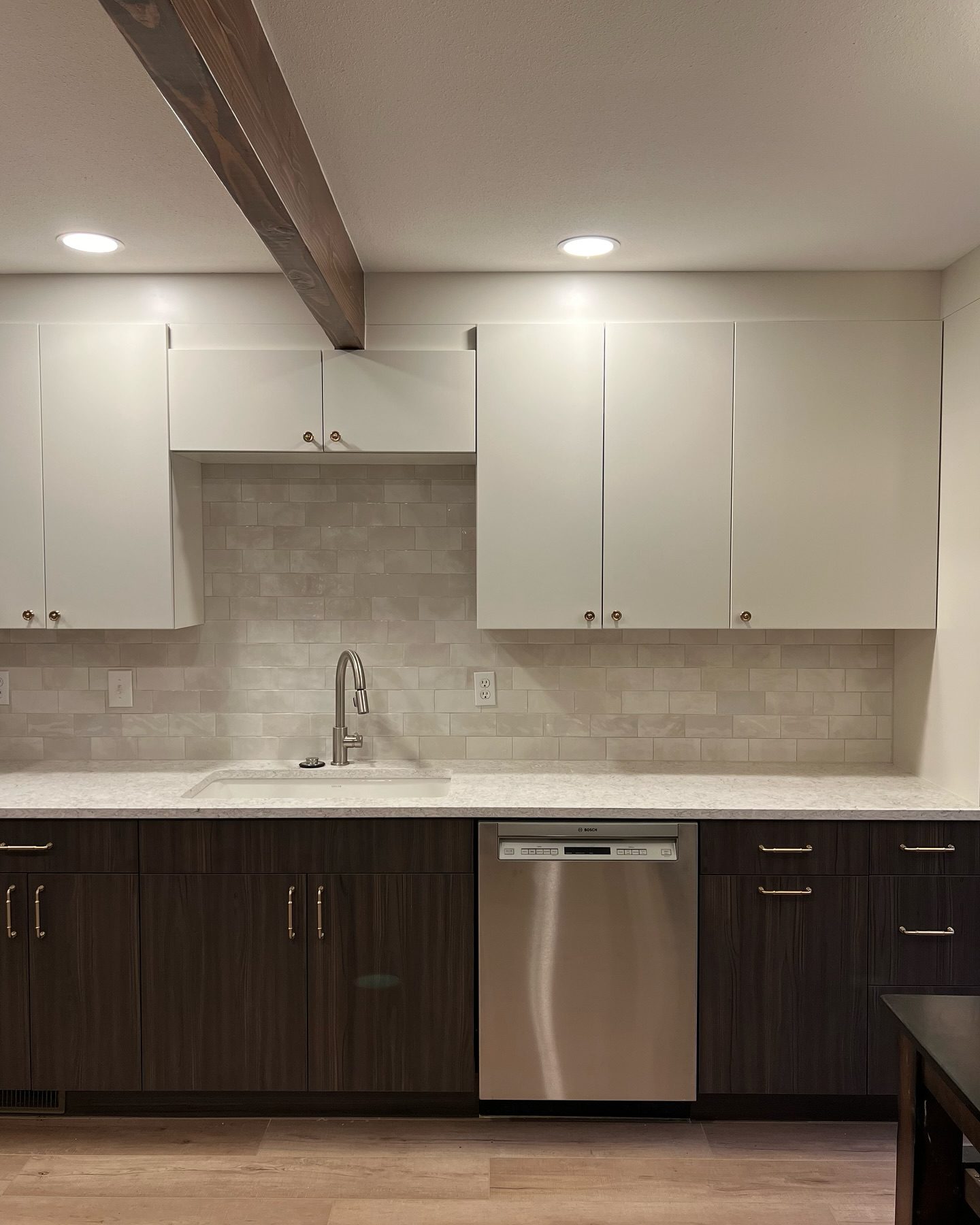 Modern kitchen showroom-style sink wall with white upper cabinets, dark wood lower cabinets and brass hardware, light marble-look quartz countertop, subway tile backsplash in neutral tones, stainless steel dishwasher and single-handle pull-down faucet under recessed ceiling lights and exposed wood beam