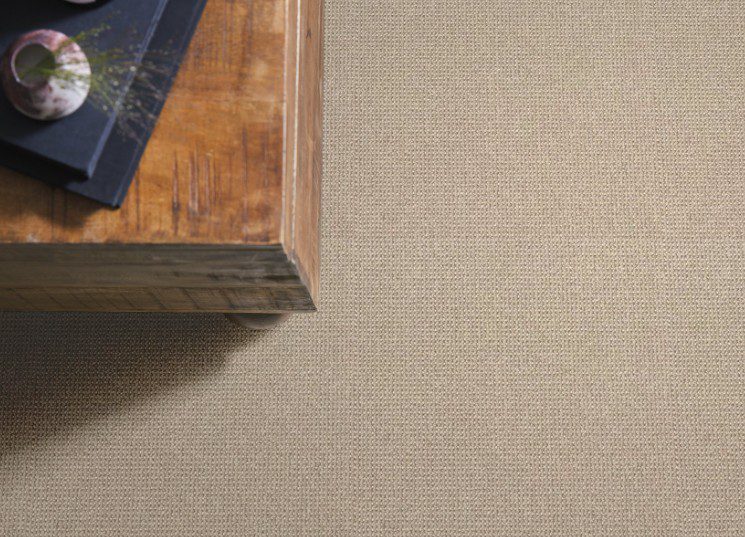 Top-down view of a rustic wooden coffee table corner on a neutral beige textured carpet, suggesting flooring and carpet options for a showroom or home design project