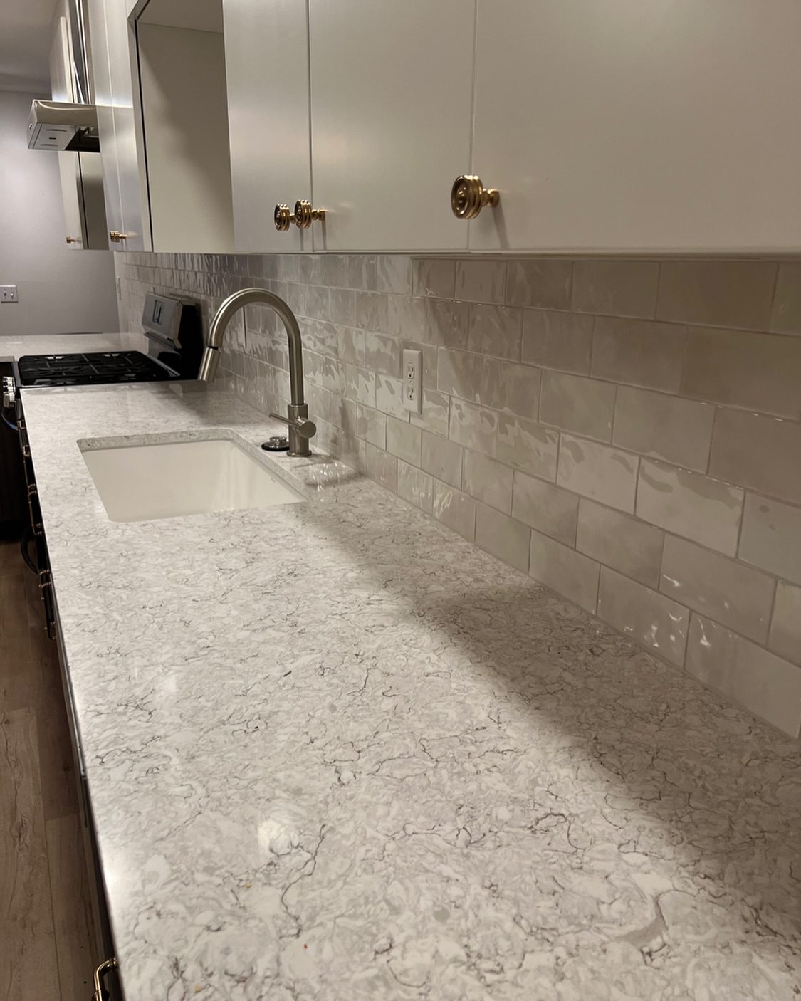 Kitchen countertop and backsplash: white quartz-look countertop with subtle gray veining, undermount sink and brushed nickel faucet, glossy subway-tile backsplash, and white upper cabinets with brass knobs — modern kitchen remodel product selection example