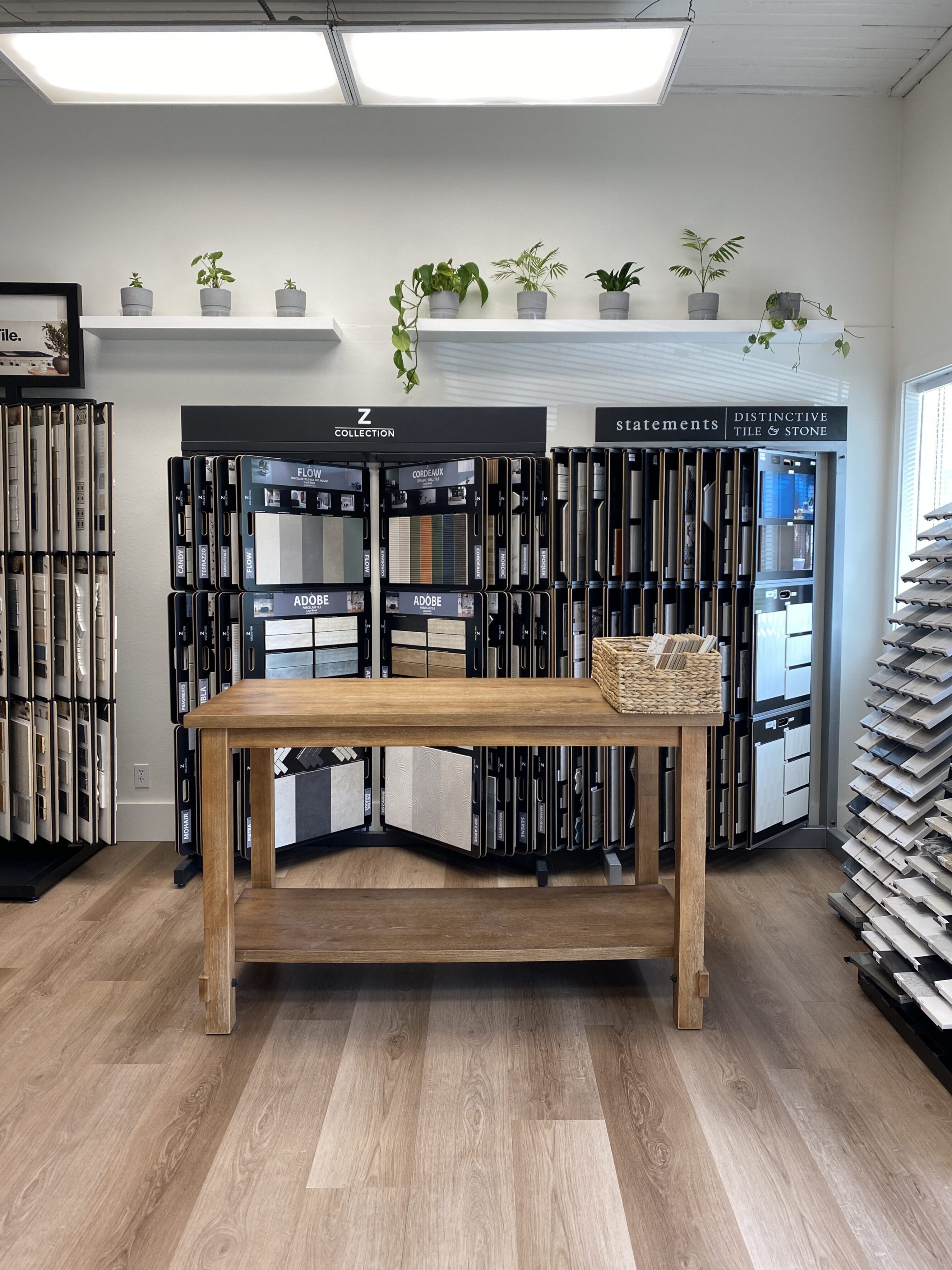 Bright showroom corner of Living By Design featuring a wooden display table on wide-plank luxury vinyl flooring in warm oak tones, surrounded by organized tile and countertop sample racks labeled "Z Collection" and "statements distinctive tile & stone," with potted plants on a white floating shelf above.