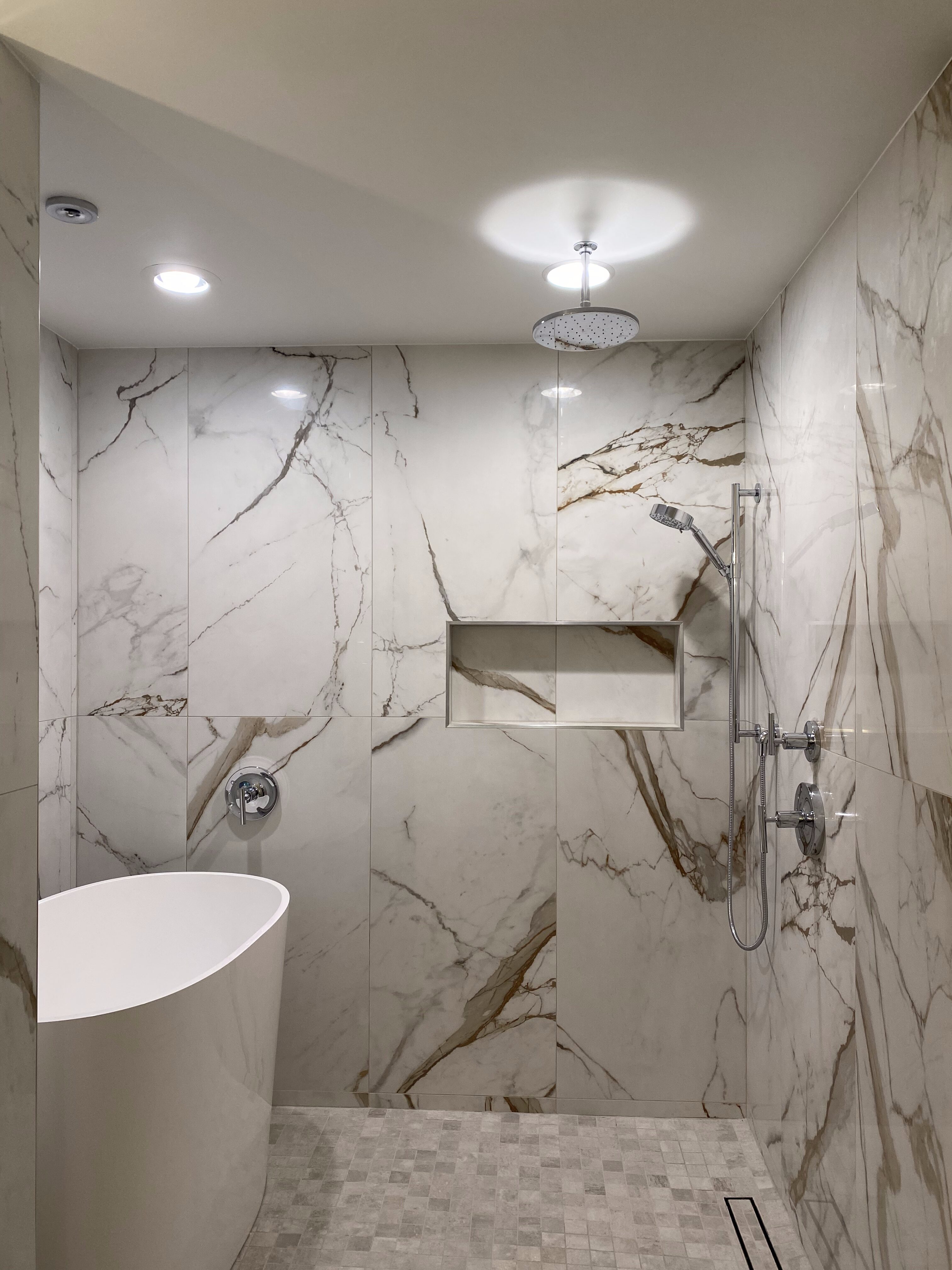 Shower and freestanding tub in a modern bathroom showroom featuring large-format marble-look porcelain tile with gold veining, built-in shower niche, rain showerhead and handheld wand, chrome fixtures, and mosaic floor tiles—luxury tile and bathroom countertop display for remodel and design services