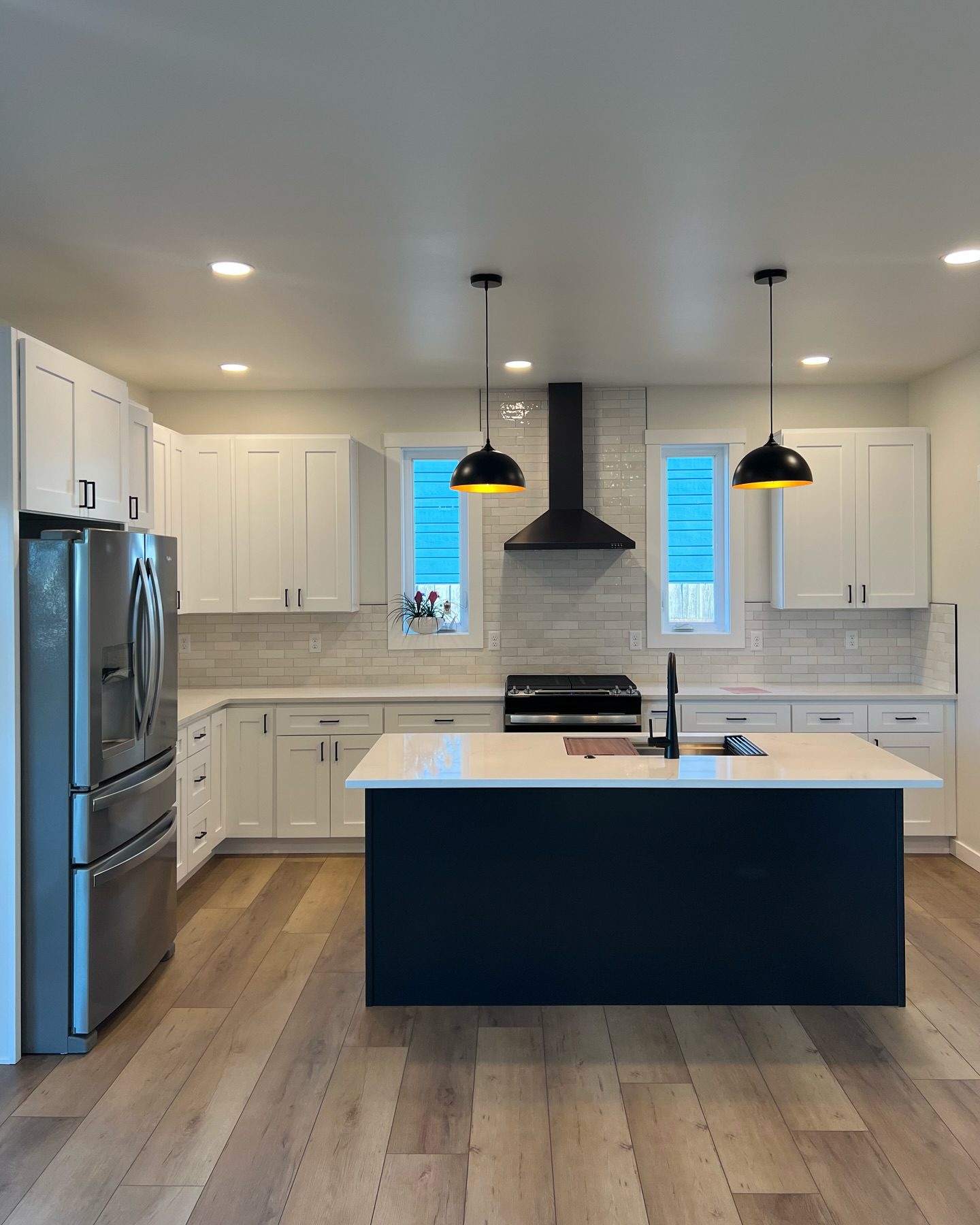 Modern kitchen remodel with white shaker cabinets, subway-tile backsplash, black island with white quartz countertop and undermount sink, stainless steel refrigerator and range, black range hood and pendant lights, and wood-look LVP flooring — showroom-style interior design and product selection example
