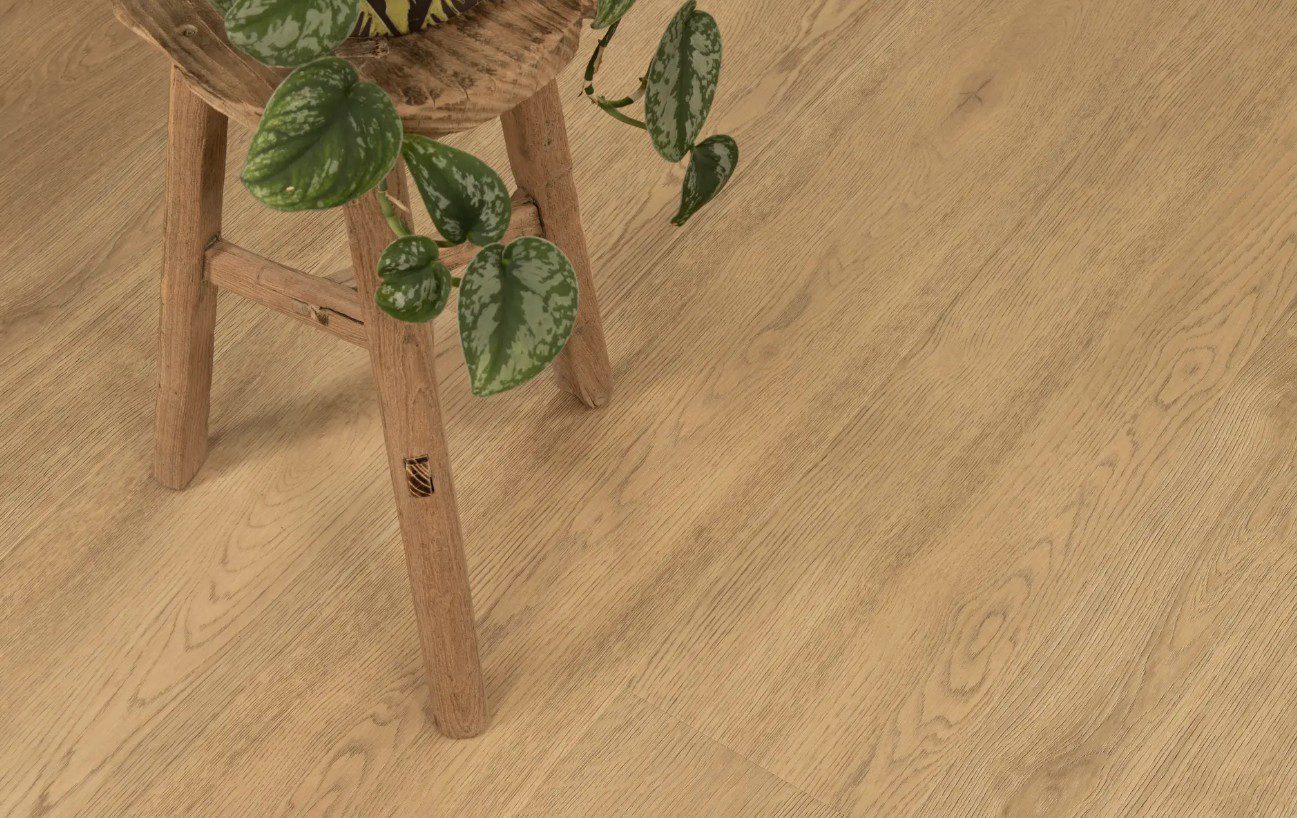 Light oak luxury vinyl plank flooring with natural wood grain texture, rustic wooden stool leg and trailing green houseplant leaves, showcasing waterproof LVP option for flooring and home design materials