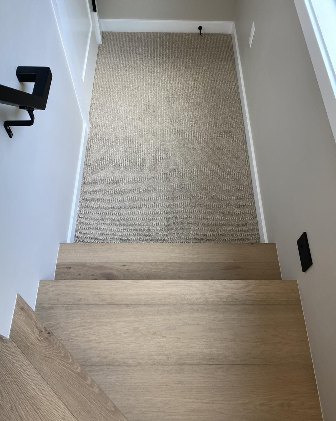 Light oak wood stair treads leading down to a small landing with neutral looped beige carpet, white baseboards, modern black square handrail and black wall outlet, showing flooring and carpet selection for a kitchen/bath or entry remodel.