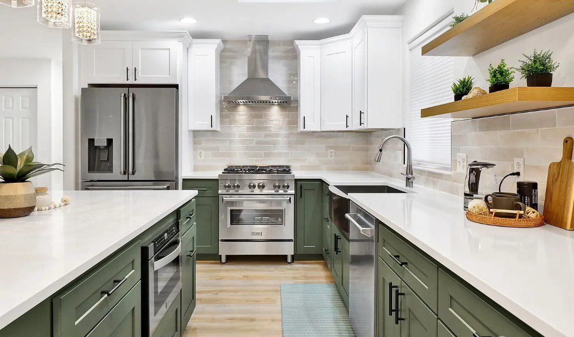 Bright modern kitchen with white quartz countertops, sage-green lower cabinets and white upper cabinets, stainless steel range and refrigerator, tile backsplash, farmhouse sink, open wood shelving with plants, and a large island — kitchen remodel, cabinetry planning, and countertop selection showcase.