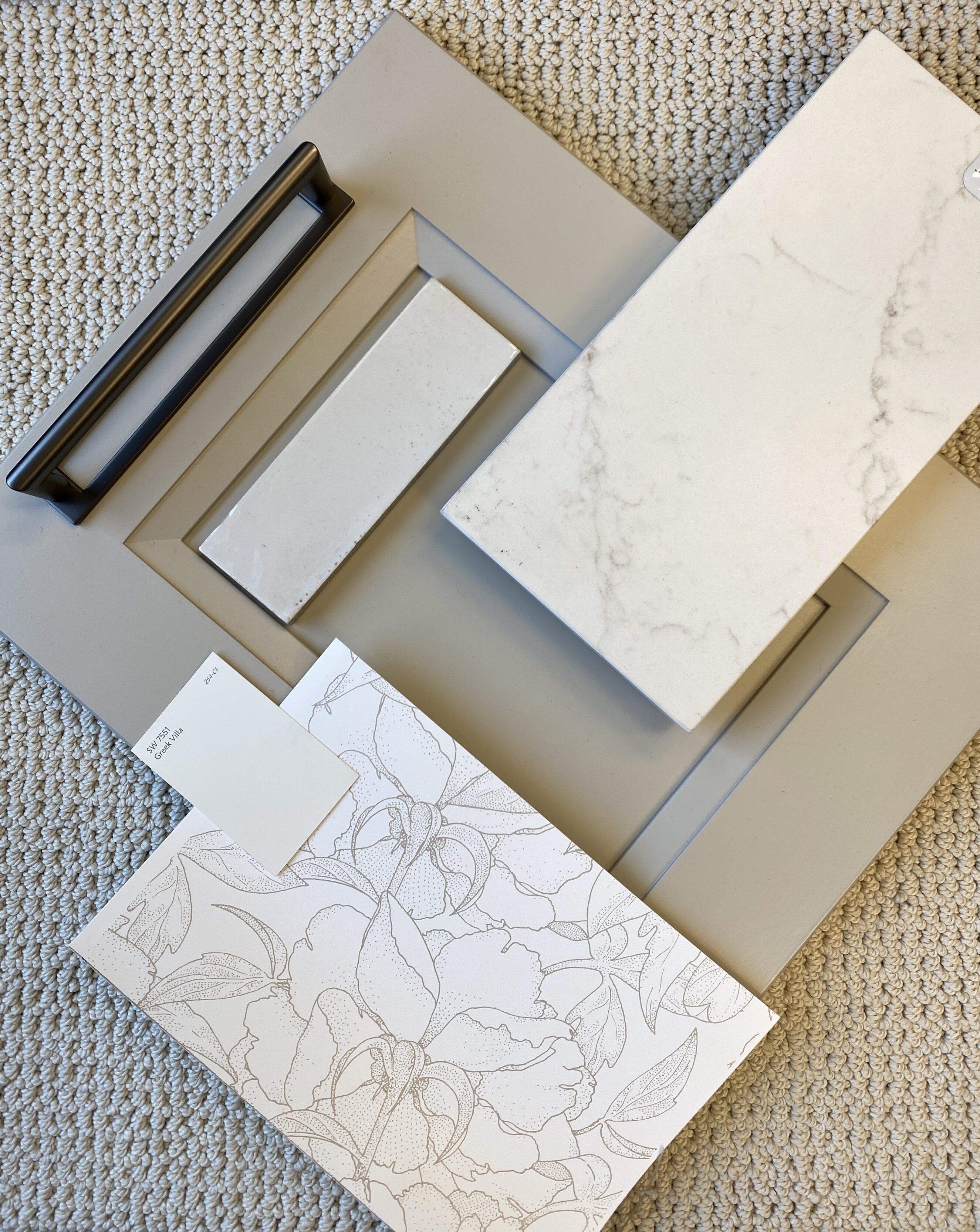 Service Page Pic Flat lay of interior design material samples for a kitchen or bath remodel: light gray shaker cabinet door with dark bar pull, marble-look white quartz countertop slab, narrow rectangular tile and a white floral wallpaper swatch on a textured neutral carpet — conveys product selection, countertop and cabinetry planning for Living by Design.