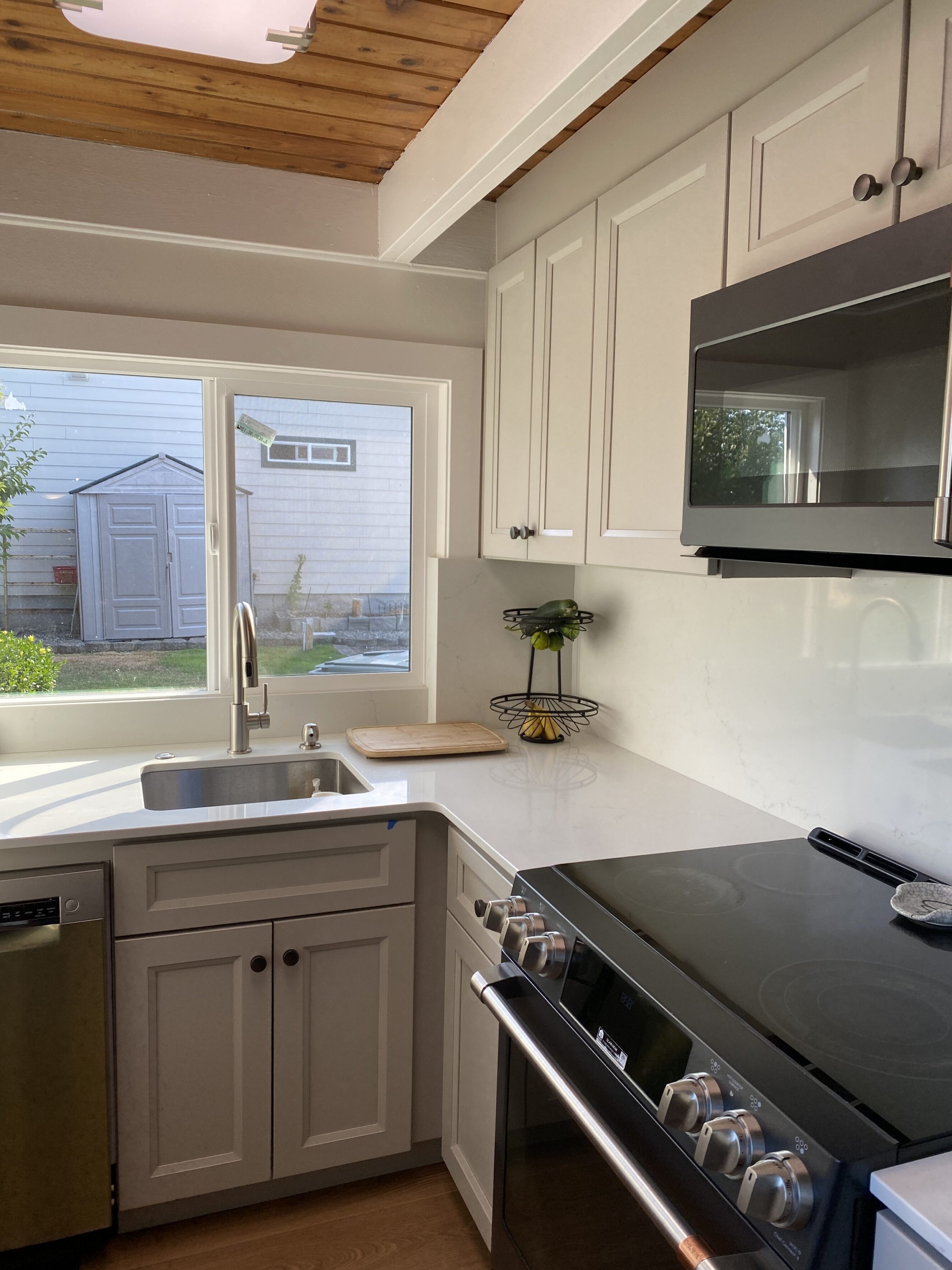 Bright remodeled kitchen corner with white quartz countertops and matching white shaker cabinets, stainless-steel undermount sink and modern faucet under a large window, stainless dishwasher to the left, black electric range and stainless microwave to the right, light wood plank ceiling and warm wood flooring, styled with a cutting board and fruit stand on the countertop — showroom-quality materials for timeless kitchen design